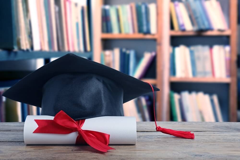 Afstudeerhoed en diploma secundair onderwijs met een rood lint voor een boekenkast.