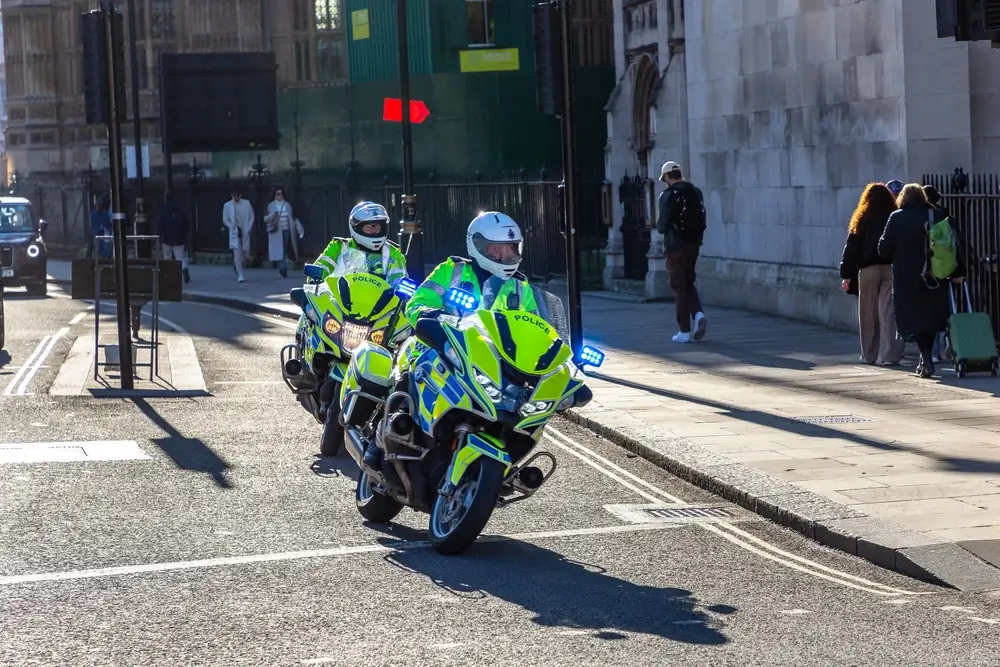 Twee politiemotorrijders met zwaailichten en sirenes tijdens een escorte en parcoursbeveiliging van een wielerwedstrijd.