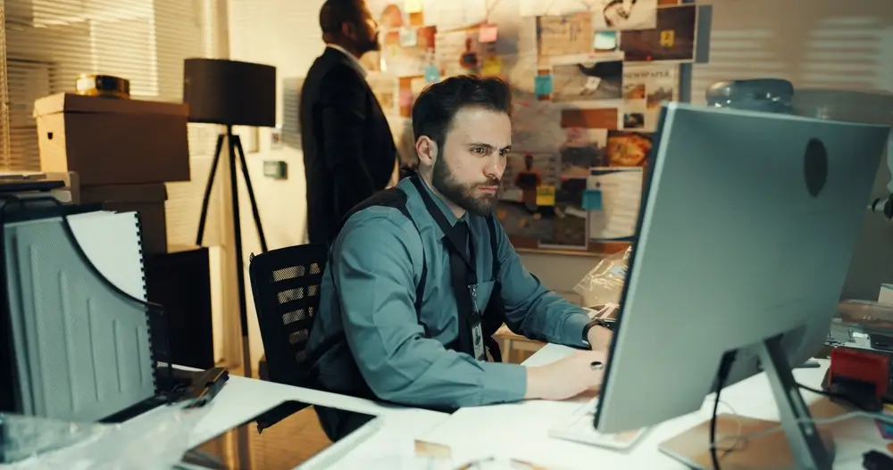 Een cyberrechercheur geconcentreerd voor zijn computerscherm, omringd door bewijsmateriaal en een onderzoeksbord.