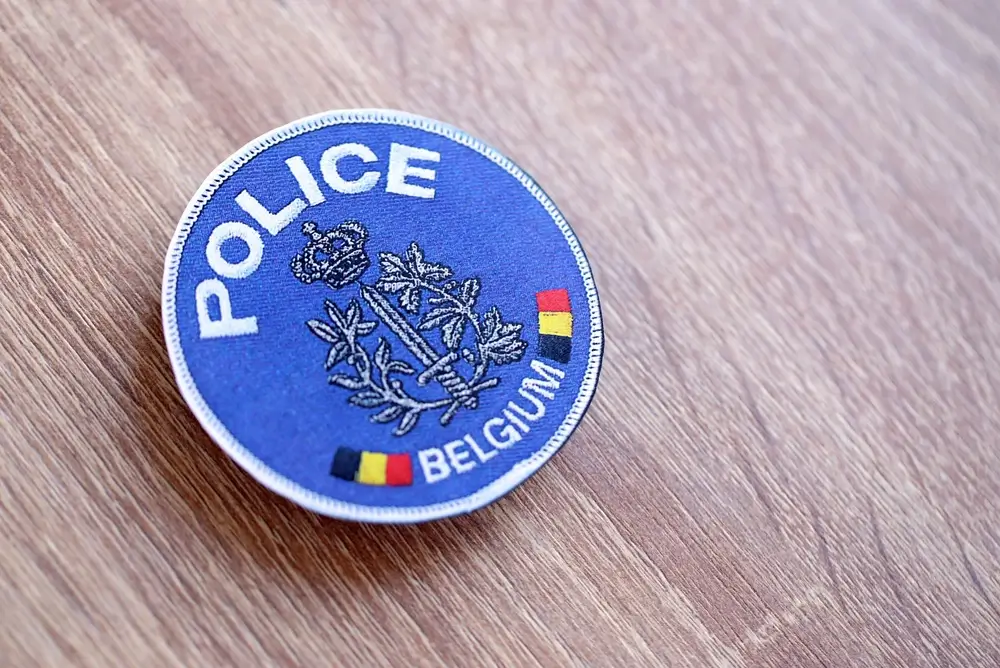 Insigne circulaire bleu de la police belge avec l'inscription Police Belgium et les couleurs nationales sur fond bois.