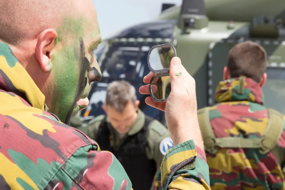 Militaire belge en tenue camouflée se maquillant avant un exercice, véhicules militaires en arrière-plan.